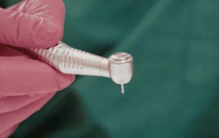 A close-up image of the head of a dental handpiece held by a hand wearing pink gloves.