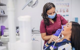 A dentist wearing a face mask works on a female patient’s teeth.